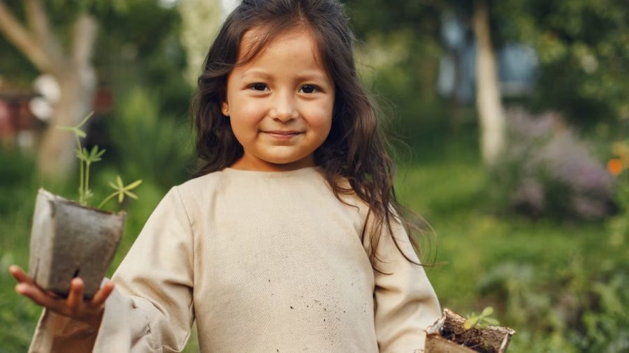 A young girl smiles while holding potted plants in a lush, sunny garden.
